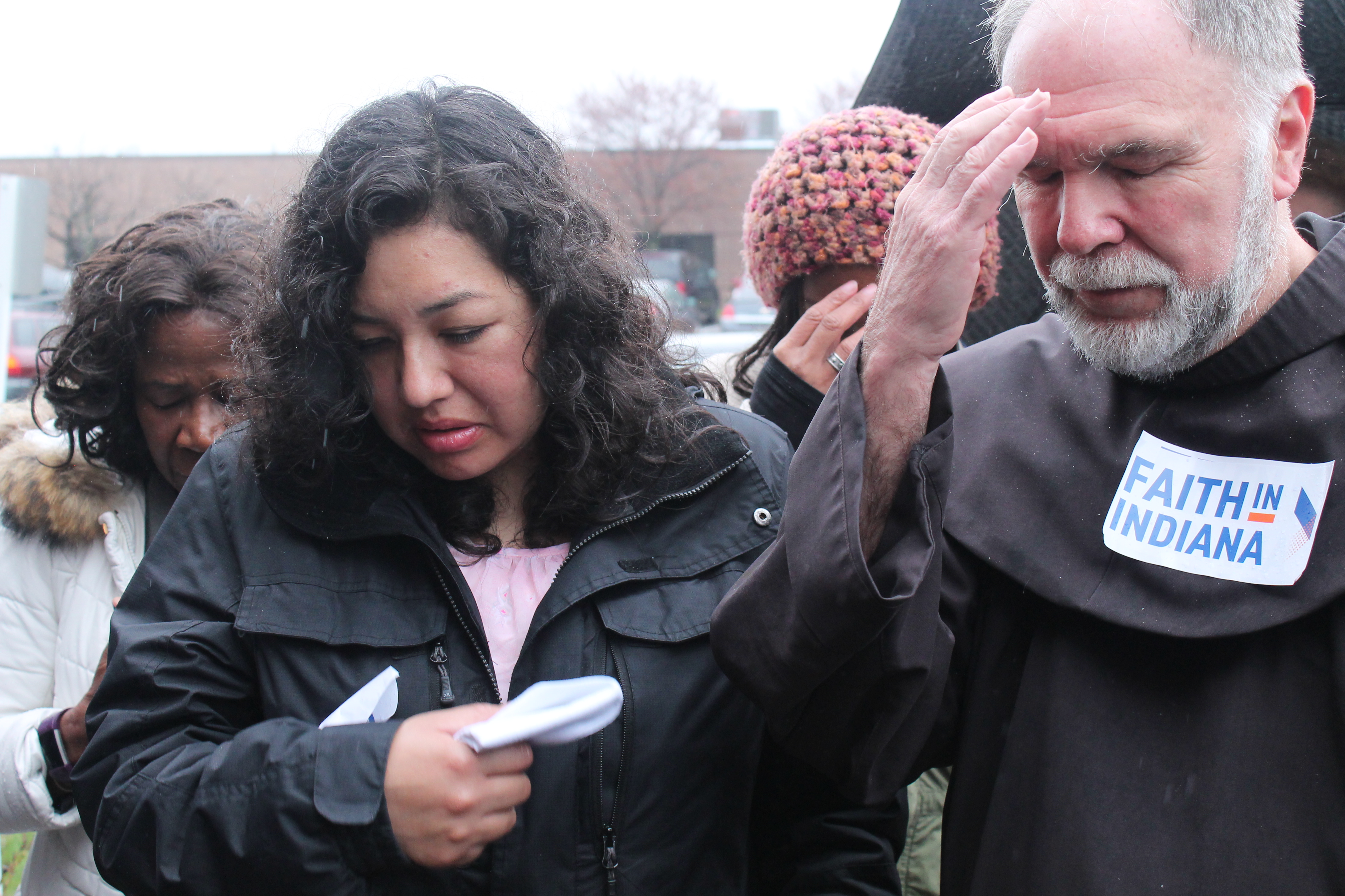 A Mexican woman, Erika Fierro, makes the sign of the cross standing next to a Catholic monk. It's raining and the monk has a 'Faith In Indiana' sticker on his robe.